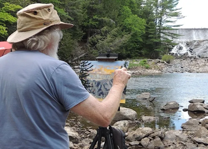 Man painting outdoors next to a rock shore and river