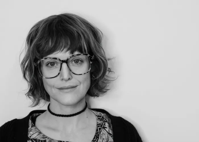 Black and white photo of performer Kelsey McNulty, wearing glasses, a necklace, and looking directly at the camera in front of a white background